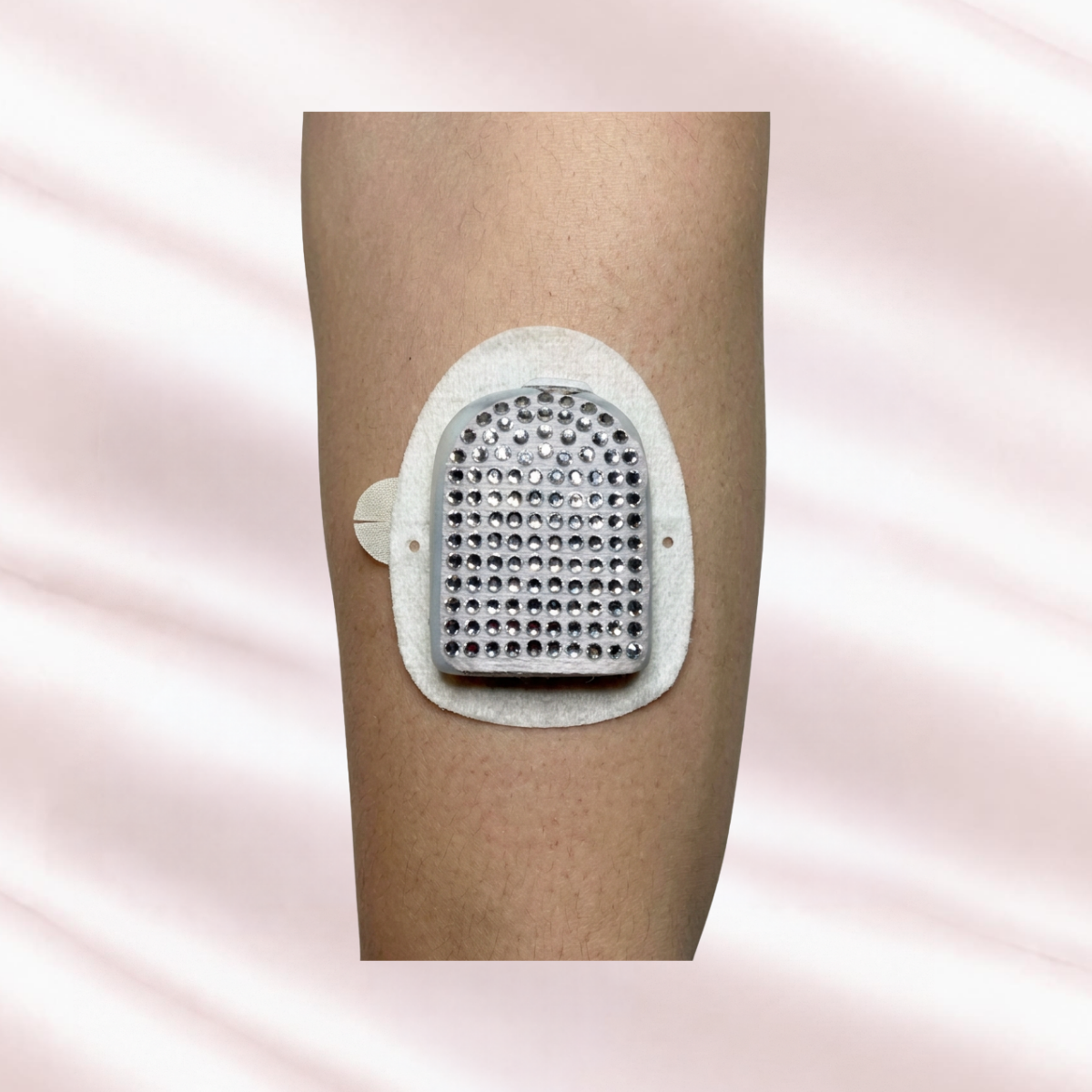 A close-up of an arm showing Omnipod Pod only Pump patch of sparkling crystal rhinestones, set against a soft, light pink satin fabric background. 